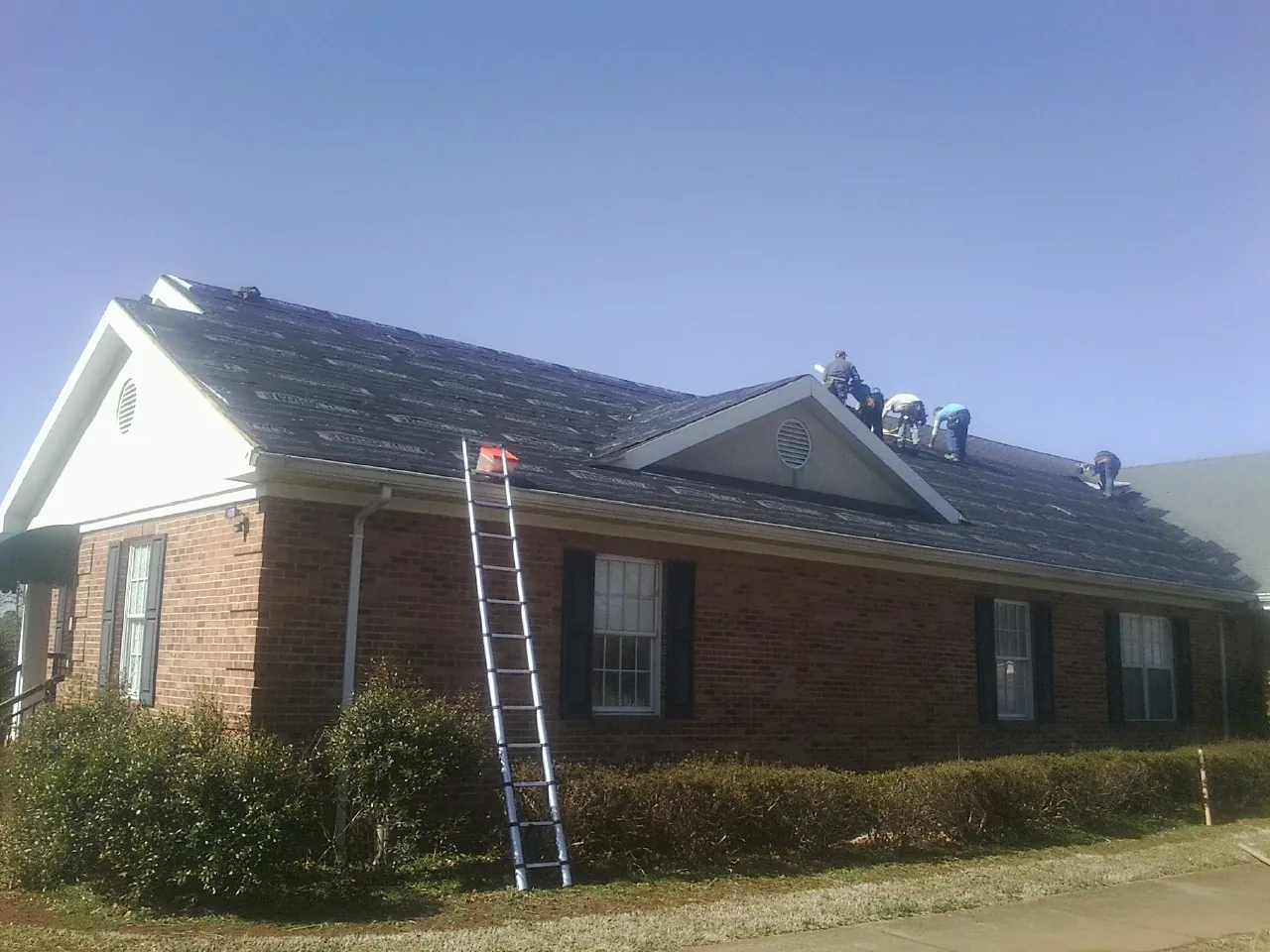 Crew tearing shingles off a brick ranch roof with a ladder set — Metal Roof Repair work by our crew in Huddleston