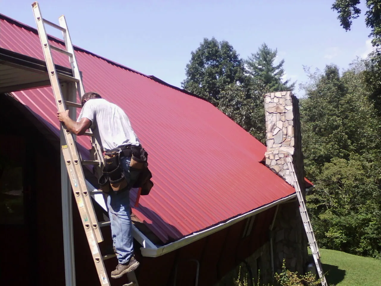 Roofer on a ladder beside a red metal roof and stone chimney — Soffit & Fascia Repair by Roofing Brothers in Huddleston